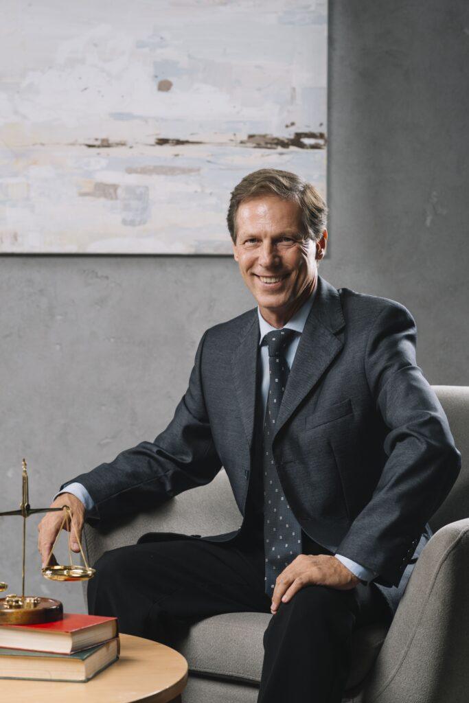 Homme en costume souriant, assis dans un bureau avec balance et livres.