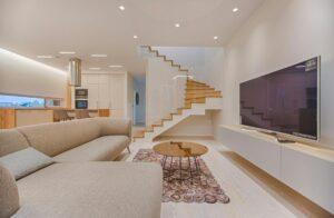 Salon moderne avec escalier en bois, canapé gris et télévision.