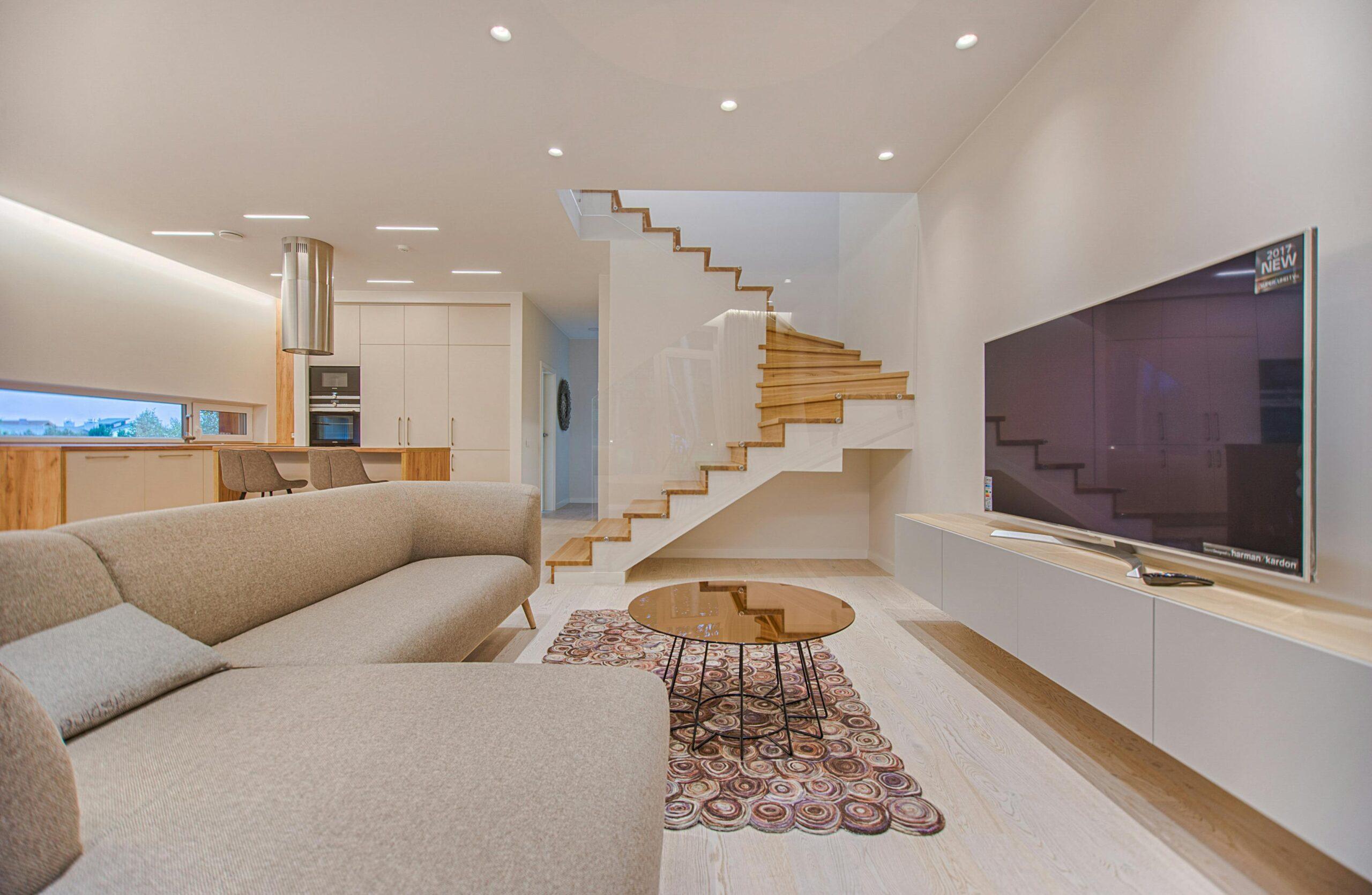 Salon moderne avec escalier en bois, canapé gris et télévision.