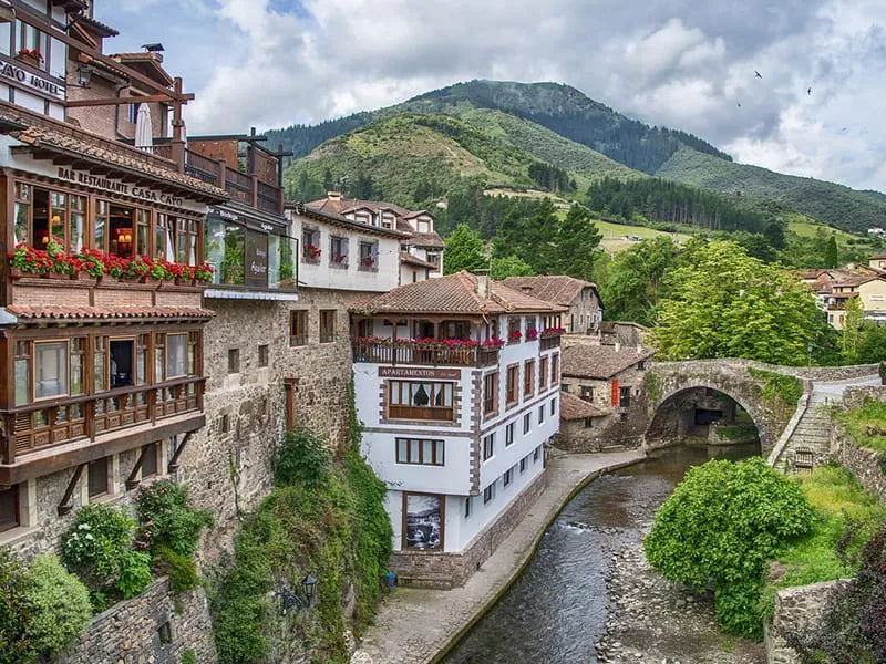 Village pittoresque avec rivière, pont en pierre et montagnes verdoyantes en arrière-plan.