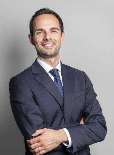 Josep Cardona Homme souriant en costume cravate sur fond gris.