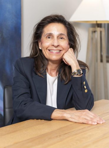 isabel alemany scaled 2 Personne souriante assise à un bureau en bois, lampe en arrière-plan.